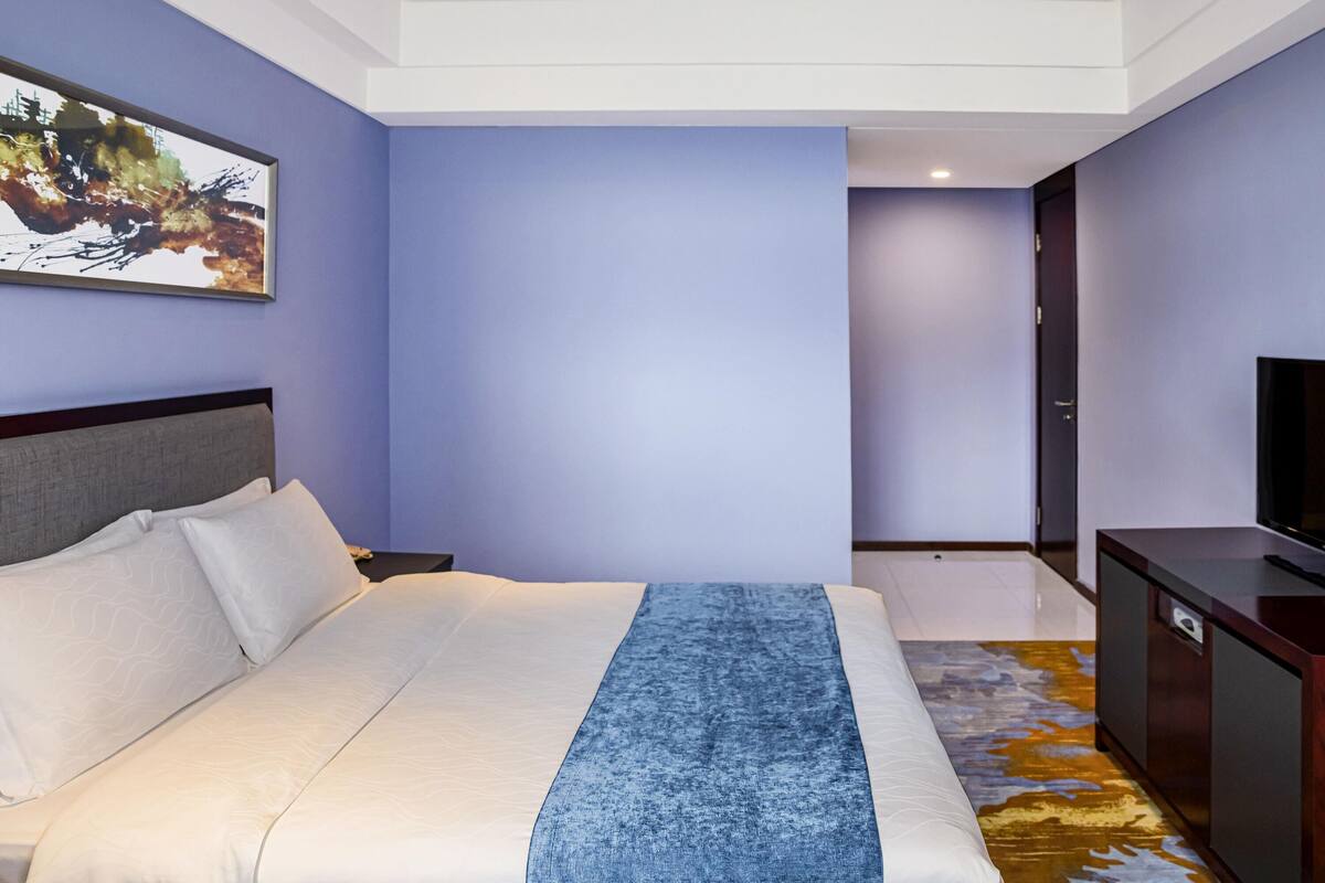 Ein Hotelzimmer mit einem großen Bett, einem Flachbildfernseher, einem Bild an der Wand und Blick auf den Flur.