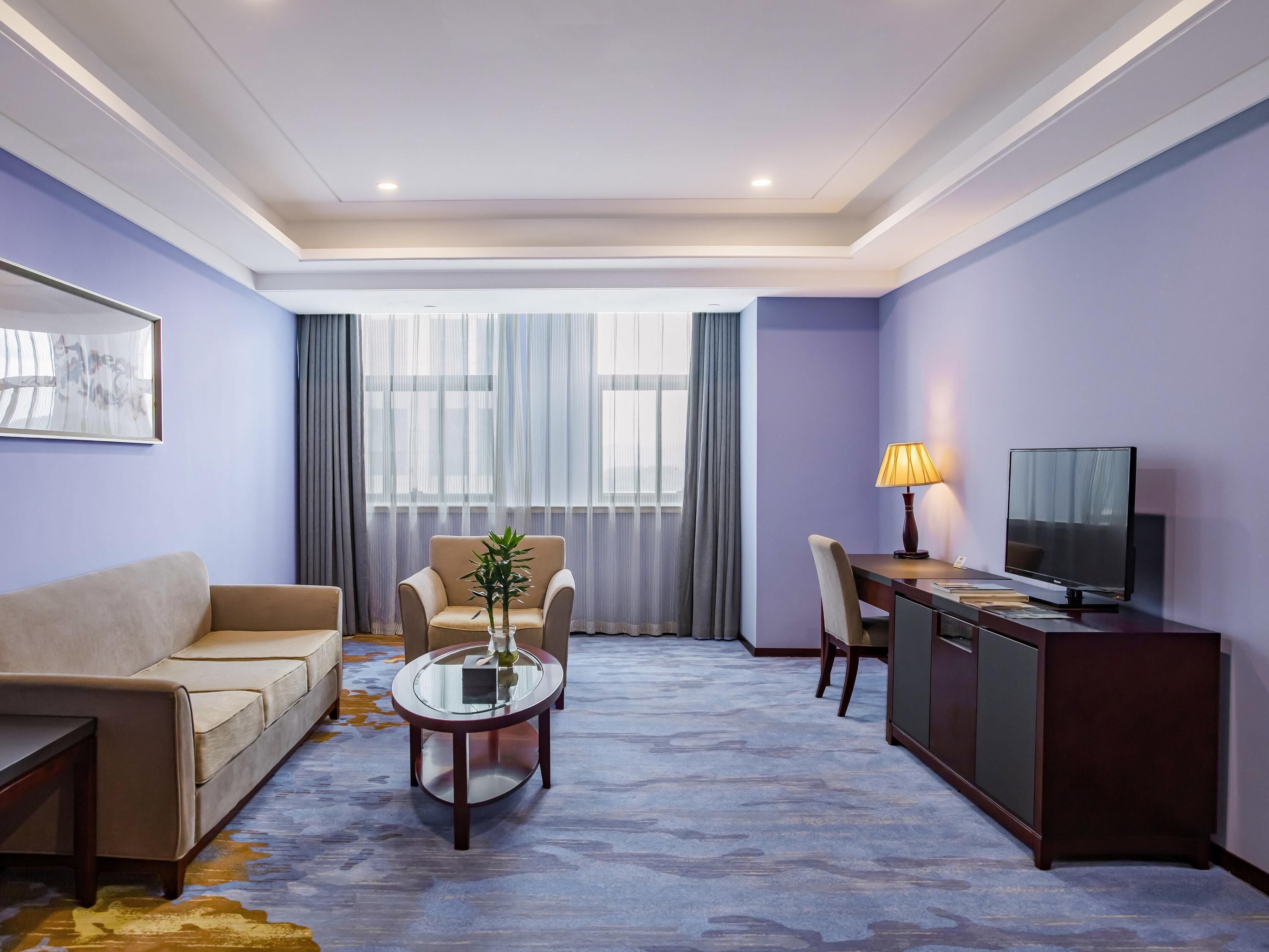 A hotel room with a sofa, a round table with a vase, a desk with a lamp, and a TV.