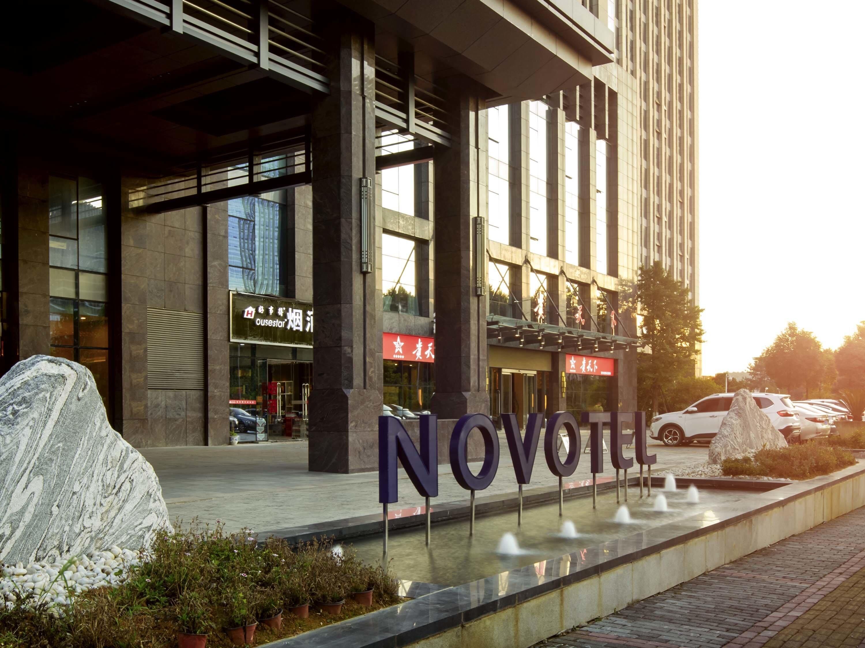 A hotel entrance with a large 'NOVOVEL' sign, a stone sculpture, and a water feature.