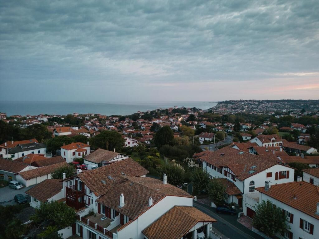 Ein Wohngebiet mit Häusern mit roten Ziegeldächern und weißen Wänden, umgeben von viel Grün und einem Blick auf das Meer in der Ferne.