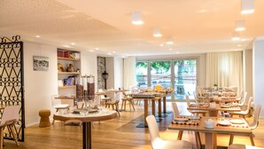 Un comedor amplio con varias mesas y sillas, una estantería con libros y objetos decorativos, y un ventanal que ofrece vistas al exterior.