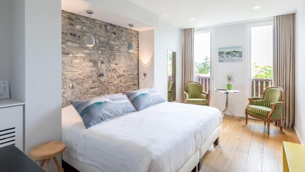 Un dormitorio con pared de piedra, una cama con ropa blanca, dos sillas, una mesita y vistas a zonas verdes.