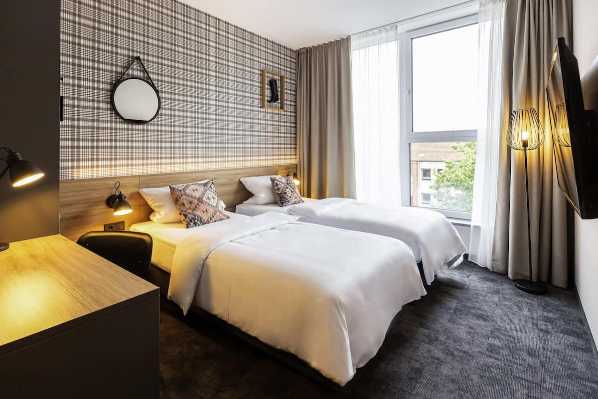 Hotellihuone, jossa on kaksi sänkyä, puinen työpöytä, peili, seinälle asennettu televisio ja ikkuna, jossa on verhot.