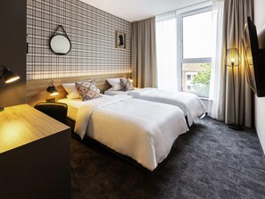 Habitación de hotel con dos camas, un escritorio de madera, un espejo, un televisor montado en la pared y una ventana con cortinas.