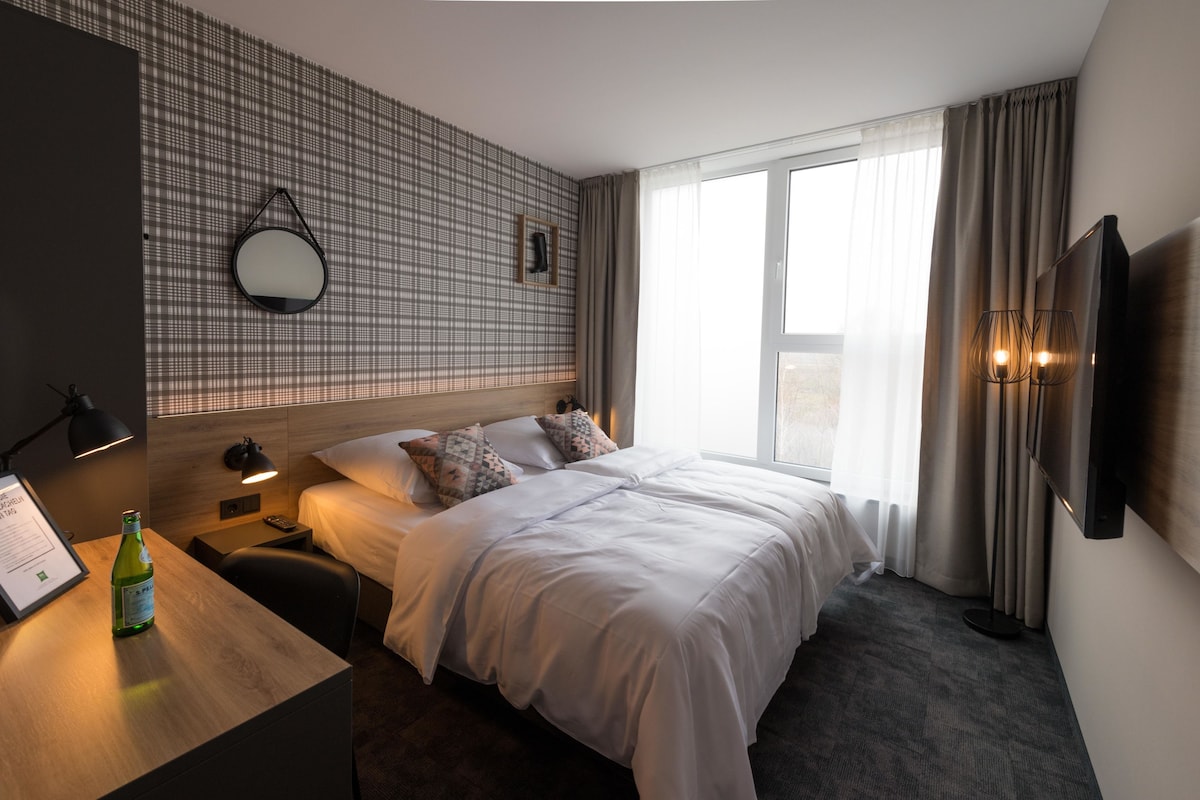 A hotel room with a large bed, a desk with a bottle, a wall-mounted TV, and a window with curtains.