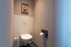 A small, modern bathroom with white subway tiles, a white toilet, a wall-mounted toilet paper holder, and a framed picture above the toilet.