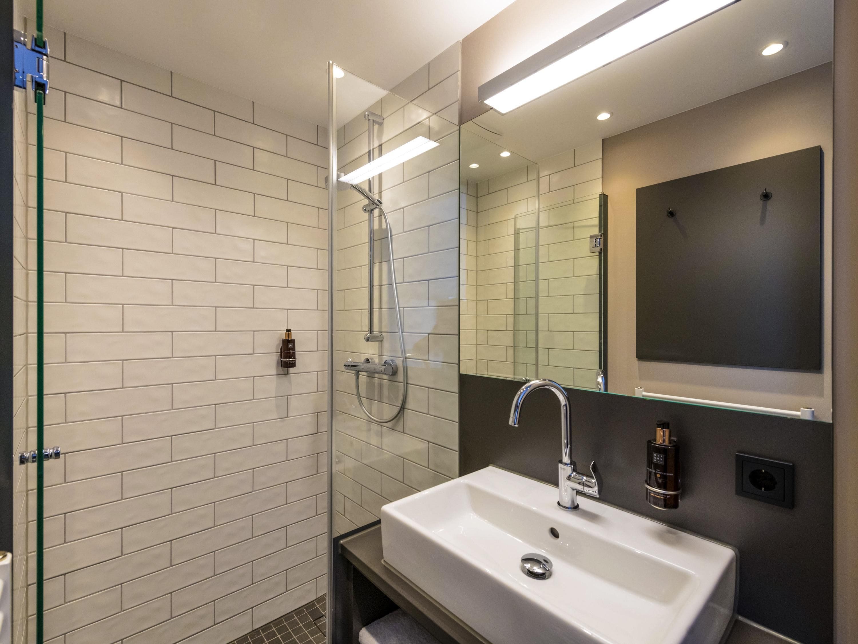 Un baño moderno con ducha acristalada, lavamanos blanco y un espejo grande.