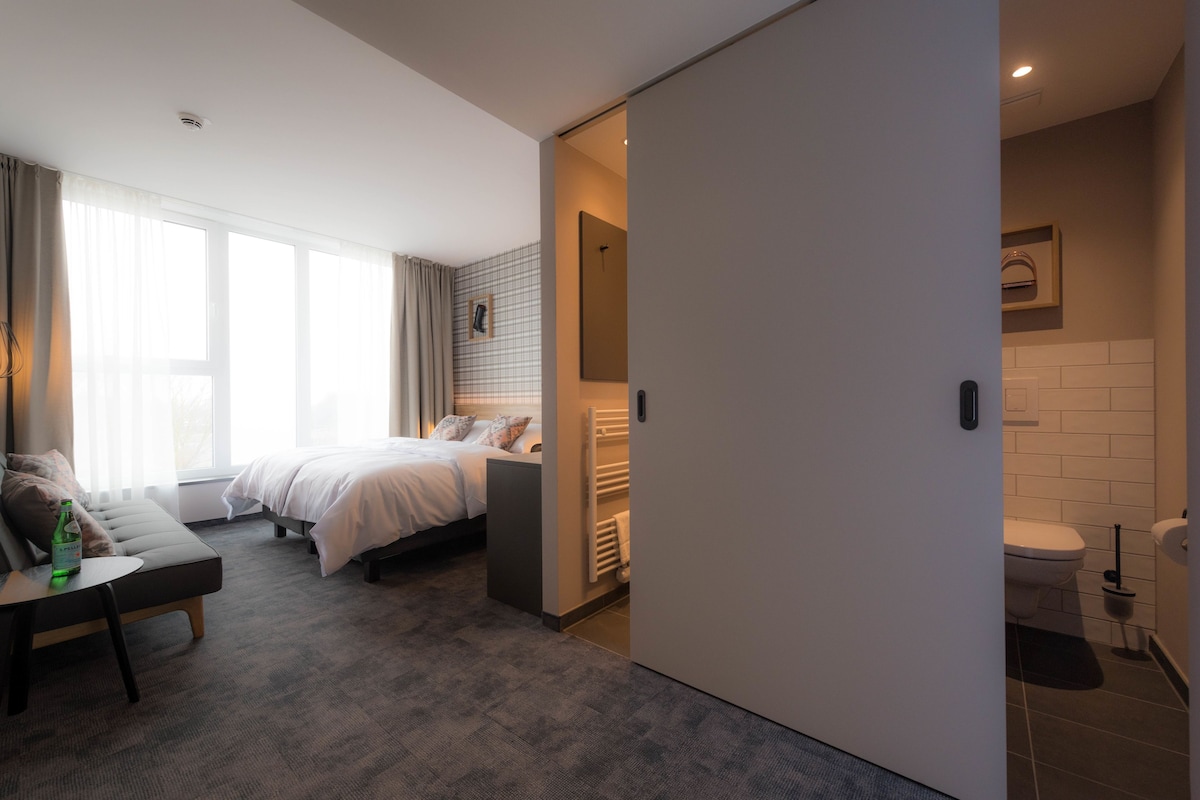 A modern hotel room with a sofa, two beds, a small table, and a view of the city.