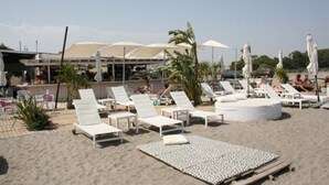 Un área de descanso frente a la playa, con tumbonas y sombrillas, una piscina con sillones y un edificio de techo curvo al fondo.