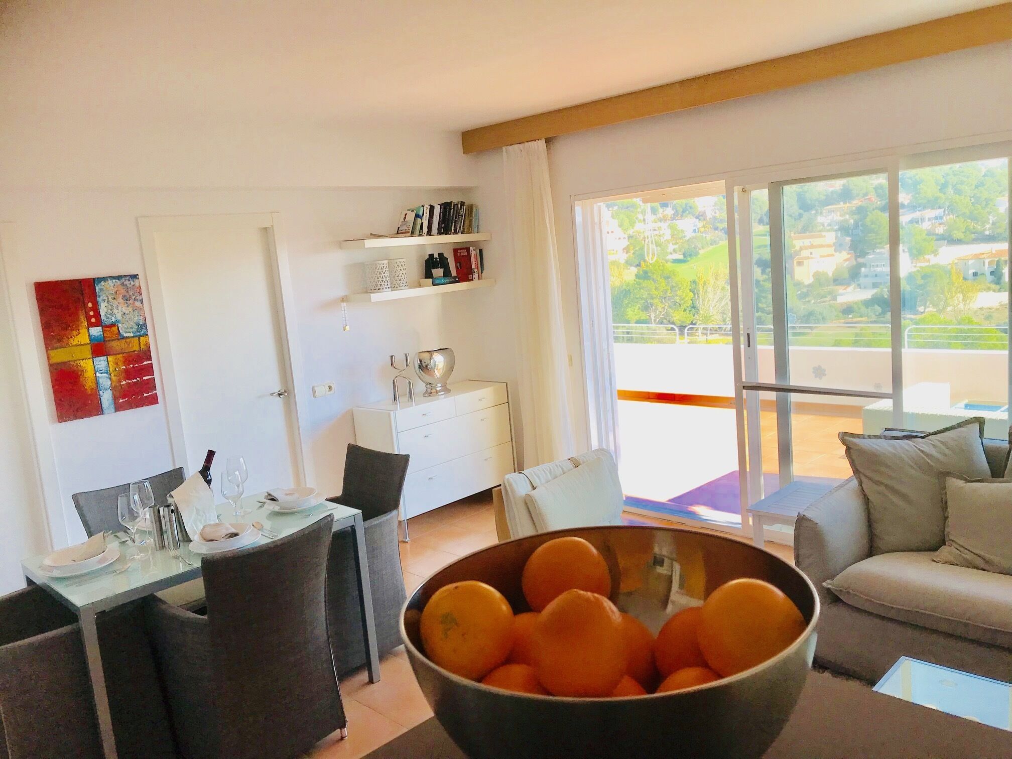 Ein modernes Wohnzimmer mit Essbereich, einem großen Fenster mit Blick ins Grüne und einer Schale Orangen auf dem Boden.
