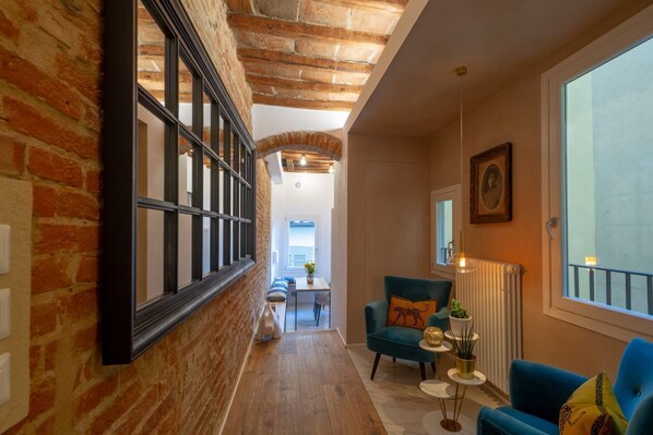A hallway with exposed brick walls, a wooden floor, a blue armchair, a small table with a plant, and a radiator.