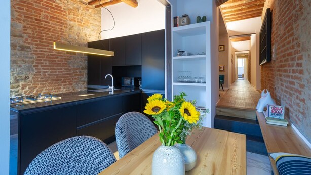 A modern kitchen with a brick wall, a wooden dining table with a vase of sunflowers, and a kitchen island with a sink and stove. - Apartments Florence - Santo Spirito Exclusive (Firenze)
