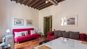 A room with a red sofa, a white table, and a wooden ceiling. There are framed pictures on the walls and a door leading to another room. - Piazza Navona Charming Apartment (Rome)
