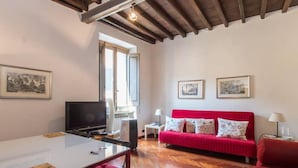 A room with a wooden ceiling, a red sofa, a TV, a dining table, and a window. - Piazza Navona Charming Apartment (Rome)