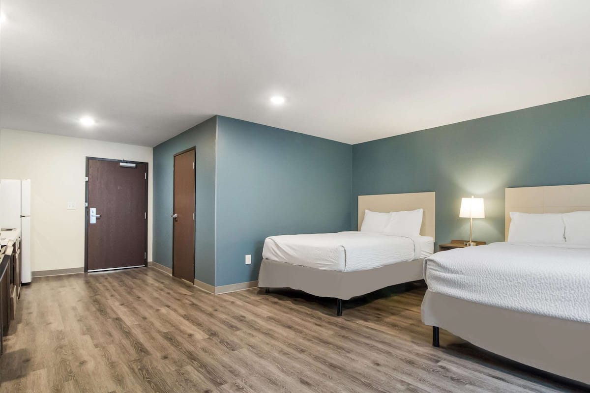 A hotel room with two beds, a wooden floor, a door, and a nightstand with a lamp.