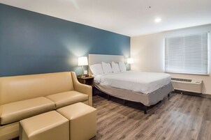 Una habitación de hotel con cama, sofá color crema, mesita de noche con lámparas y ventana con persianas.