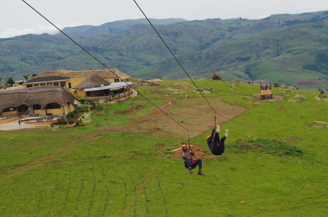 Paisaje rural con una construcción de techo de paja, un sendero de tierra y una persona en un columpio.