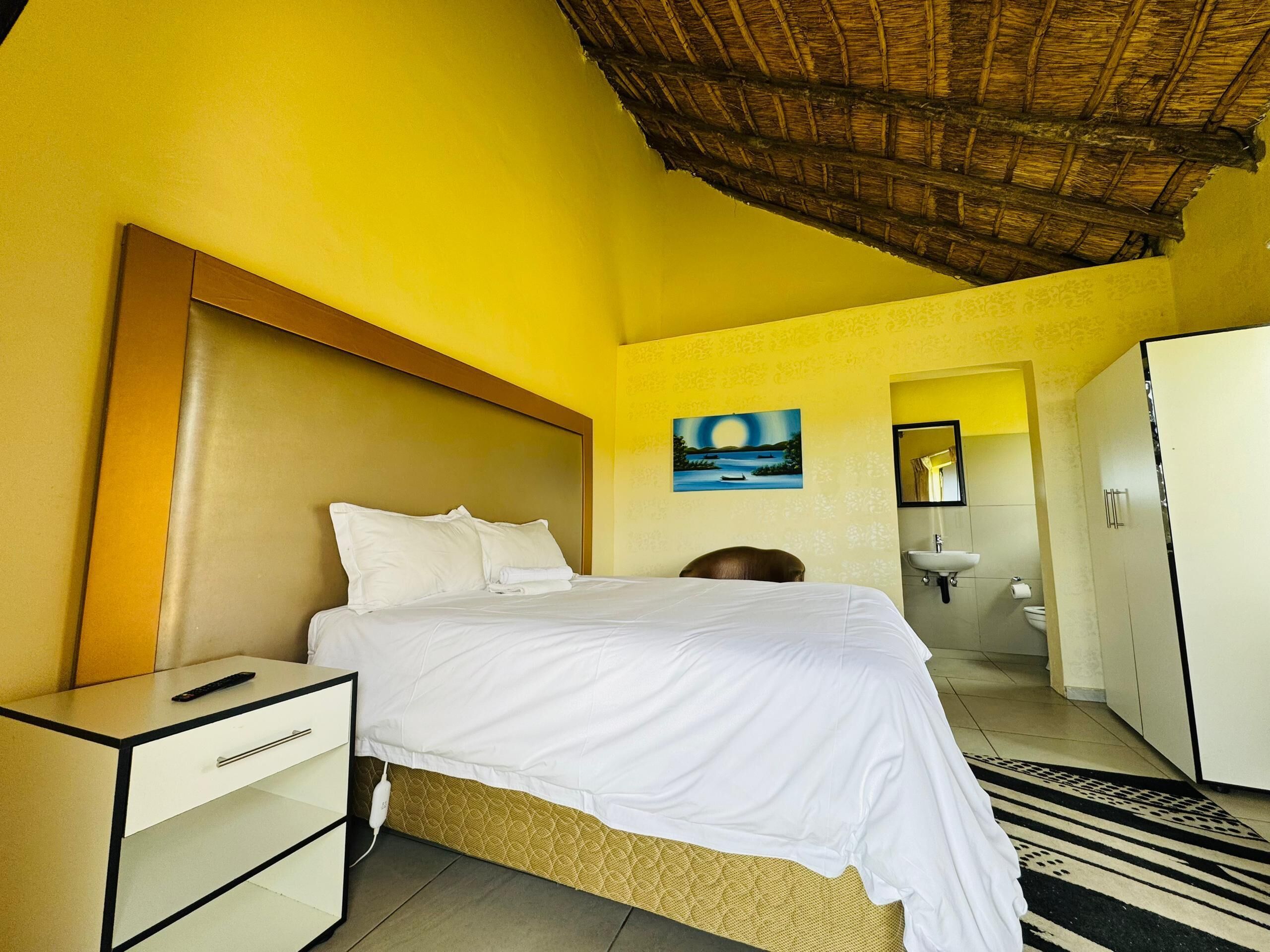 Una habitación de hotel con cama, mesita de noche, armario, baño con lavamanos e inodoro, y un cuadro en la pared.