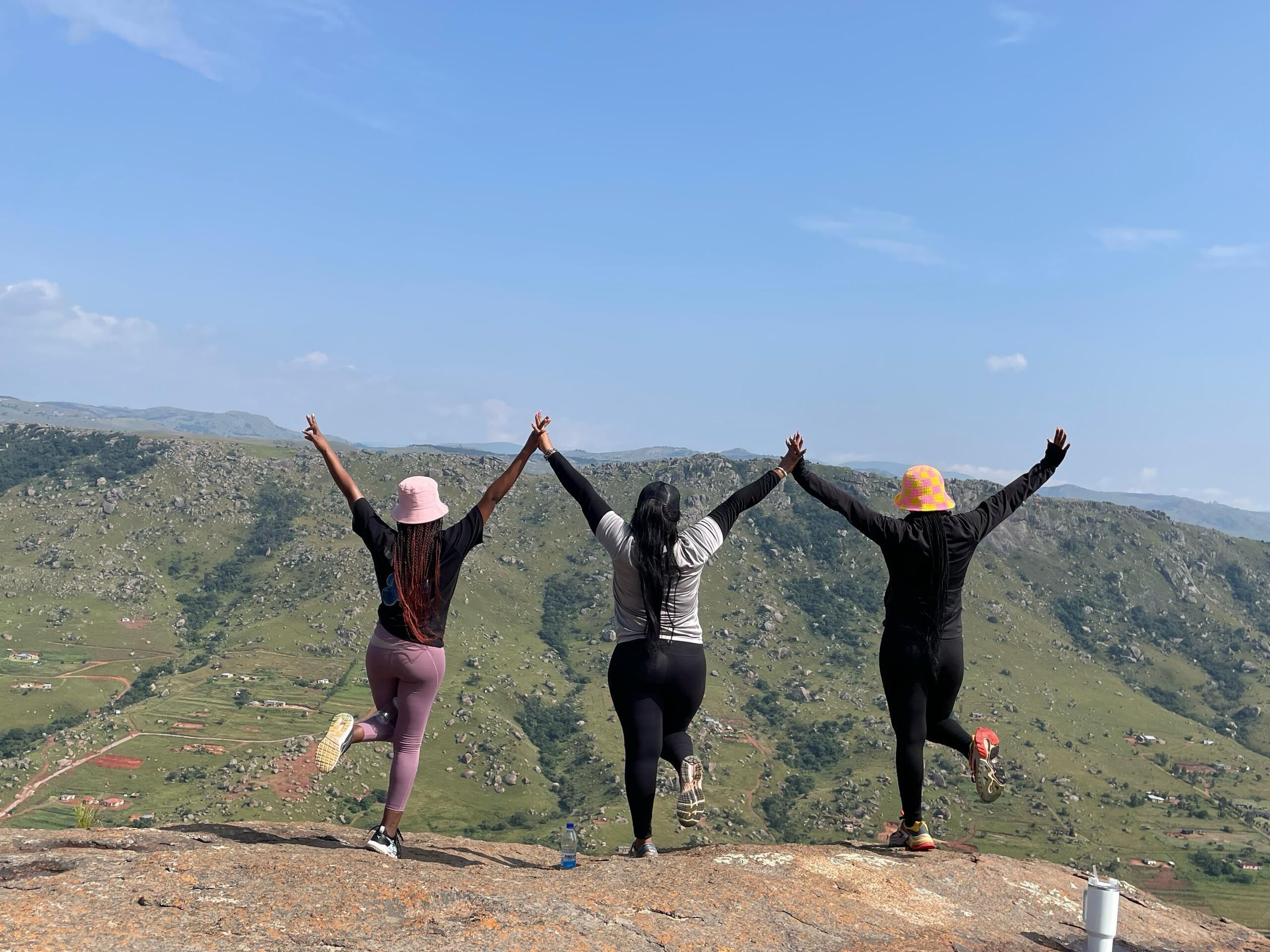 Tres personas en un saliente rocoso, con los brazos levantados, contemplando un paisaje montañoso.