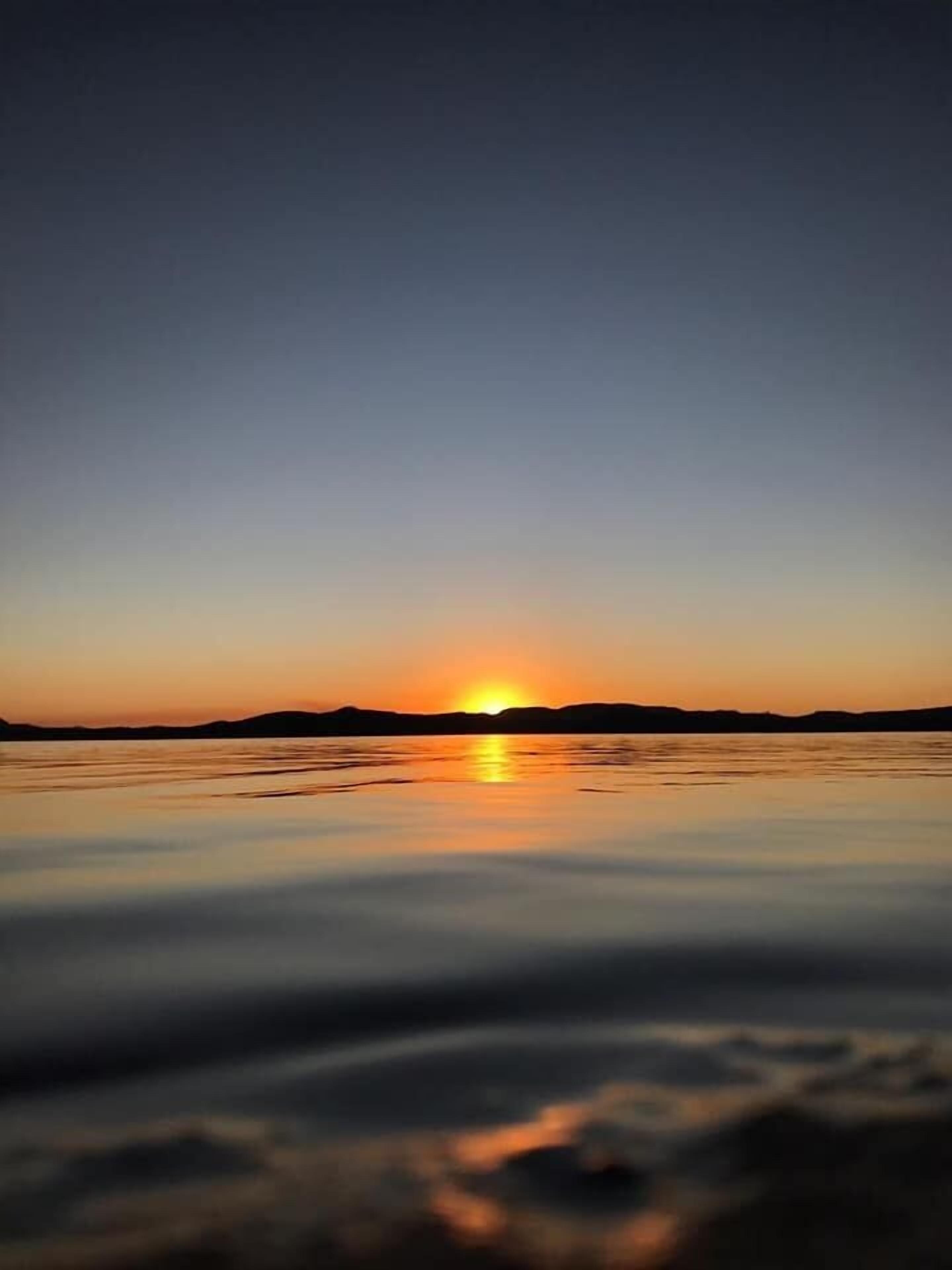 Un coucher de soleil sur une étendue d’eau calme, avec la silhouette de collines lointaines.