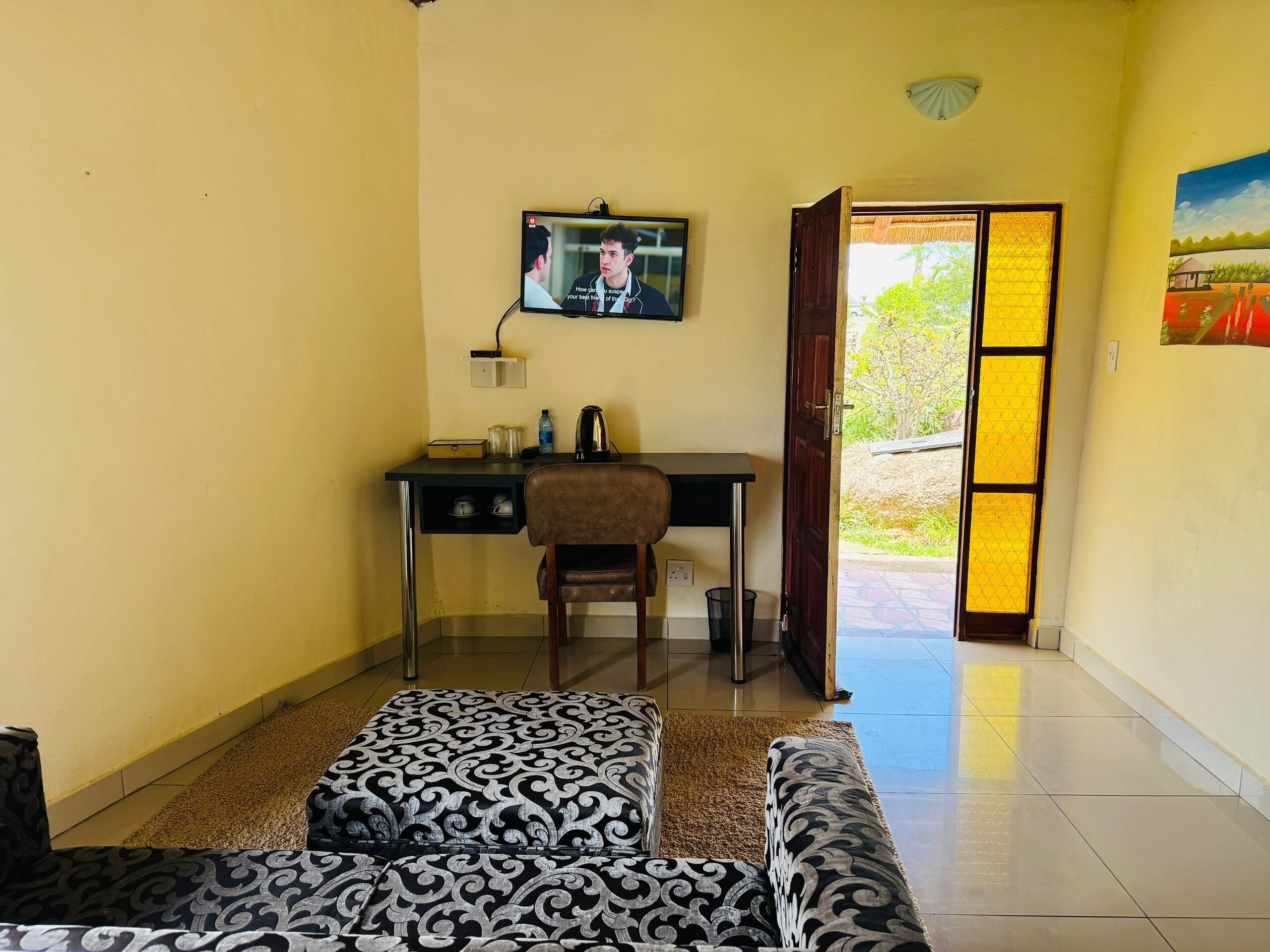 A room with a desk, chair, and a television mounted on the wall. There is a door leading outside and a window with a view of greenery.