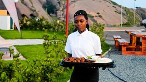A person holding a tray with food and a drink, standing in an outdoor area with a grassy lawn, a wooden bench, and a building in the background.
