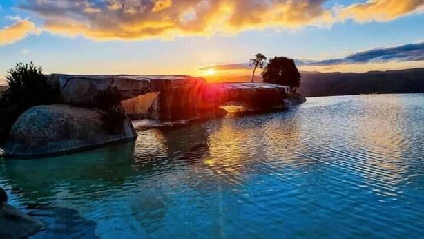 A serene lakeside view with a sunset, calm water, and rocky formations.