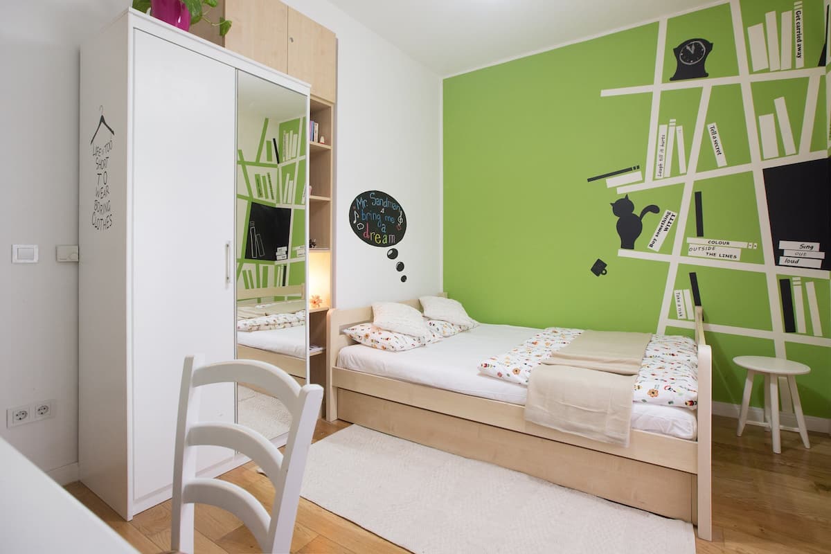 A bedroom with a bed, a wardrobe, a chair, and a wall with a bookshelf and decorative items.