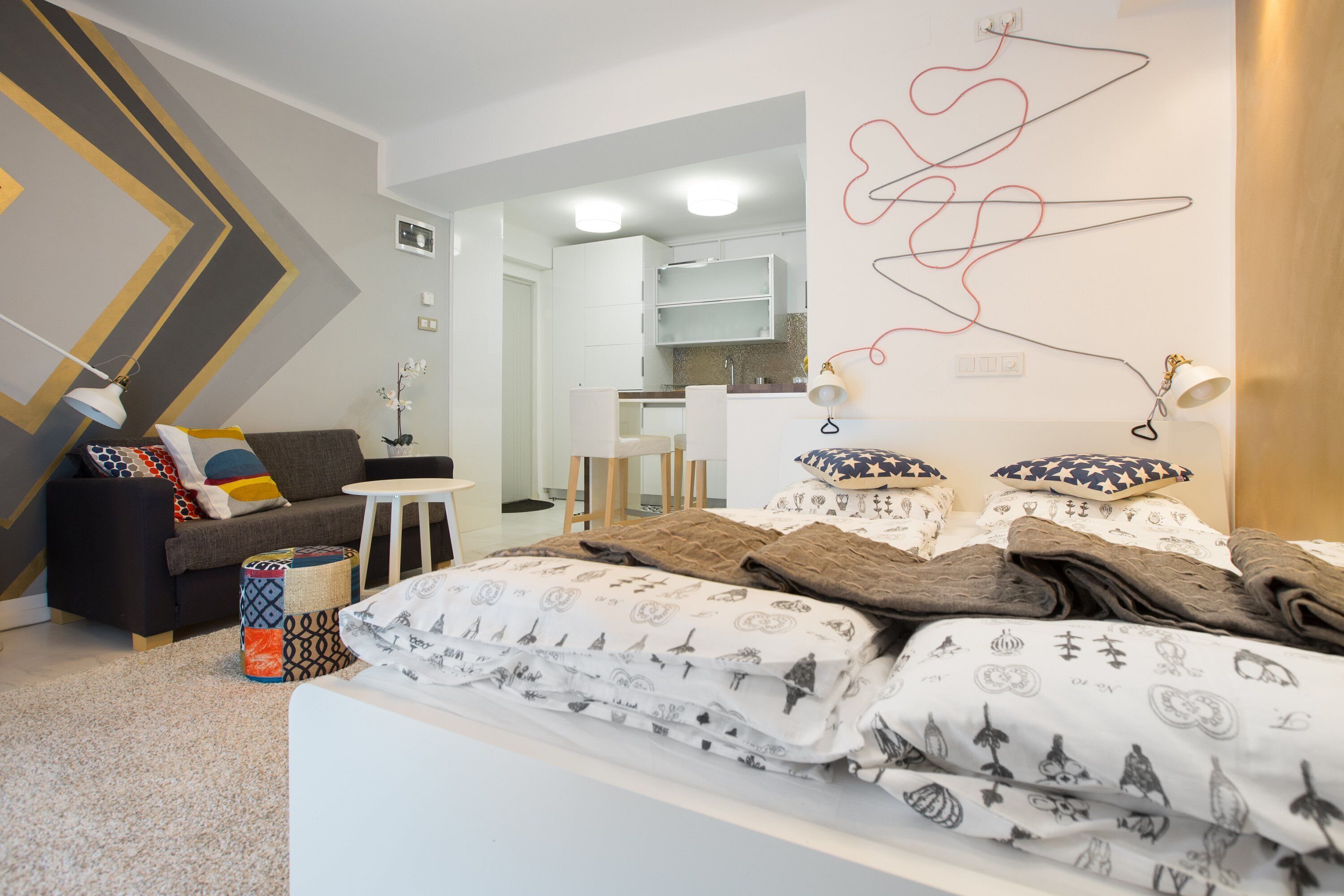 A modern bedroom with a bed, a sofa, a small table, and a kitchen area visible through an open door.