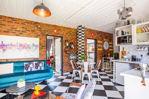 A living room with a blue sofa, a dining area with white chairs, a kitchen with white cabinets, and a brick wall. - Retro-Vintage New Yorker Apartment in Centurion (Centurion)