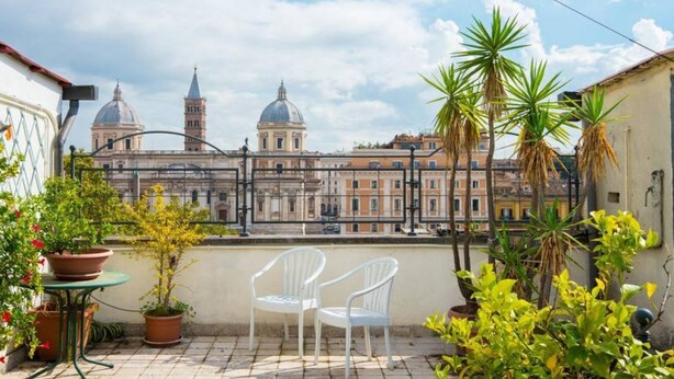 A terrace with potted plants, a table, and two chairs, overlooking a historic building with domed structures. - Opera Luxury Terrace (Rome)