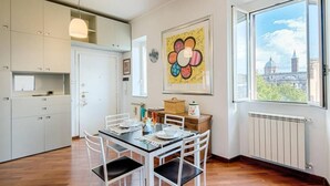 A modern kitchen with white cabinets, a dining table with chairs, a radiator, and a large window with a view of a cityscape. - Opera Luxury Terrace (Rome)