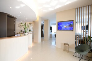 A modern reception area with a curved reception desk, a flat-screen TV mounted on the wall, and a seating area with a chair and a small table. - Albergo Grappolo D'oro (Montebelluna)
