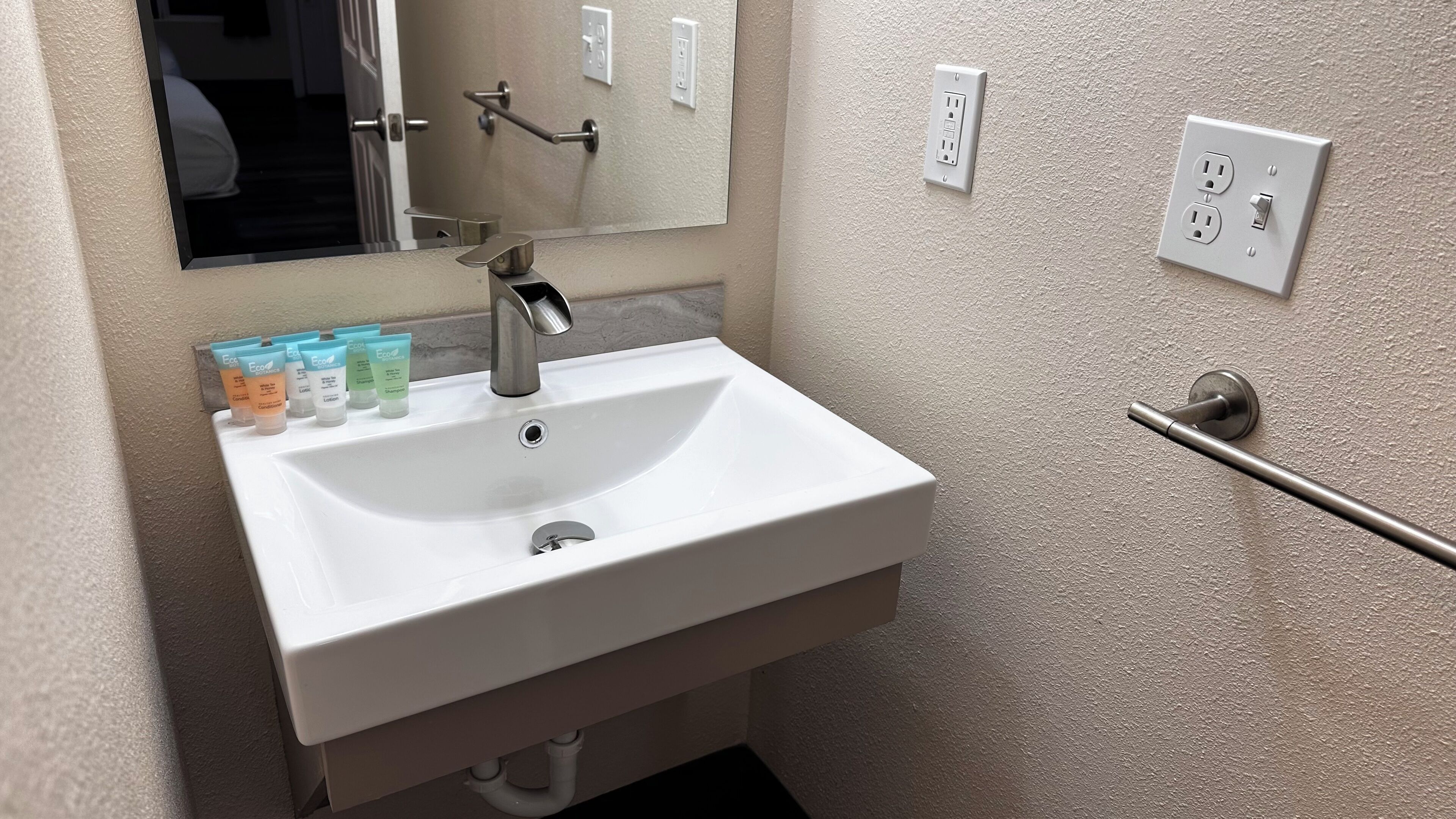 A white bathroom sink with a mirror above it, a towel rack, and a handrail. There are several toiletries on the sink counter.