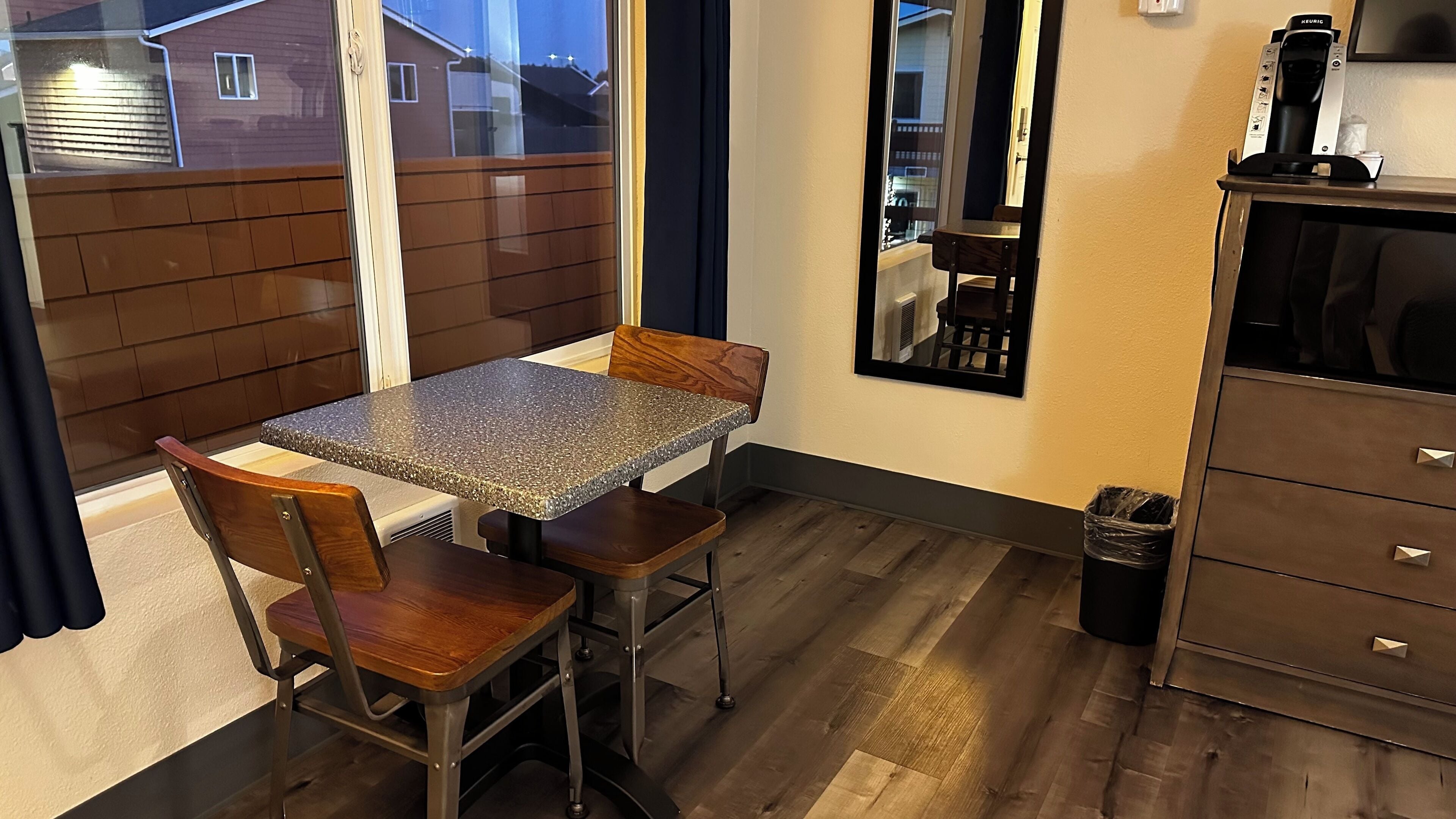 Una pequeña cocina con encimera de granito, sillas y mesa de madera, un cubo de basura y un armario con cajones.