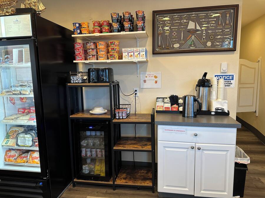 A small kitchenette area with a refrigerator, a coffee machine, and a microwave. There are shelves above with various food items and a framed picture on the wall.