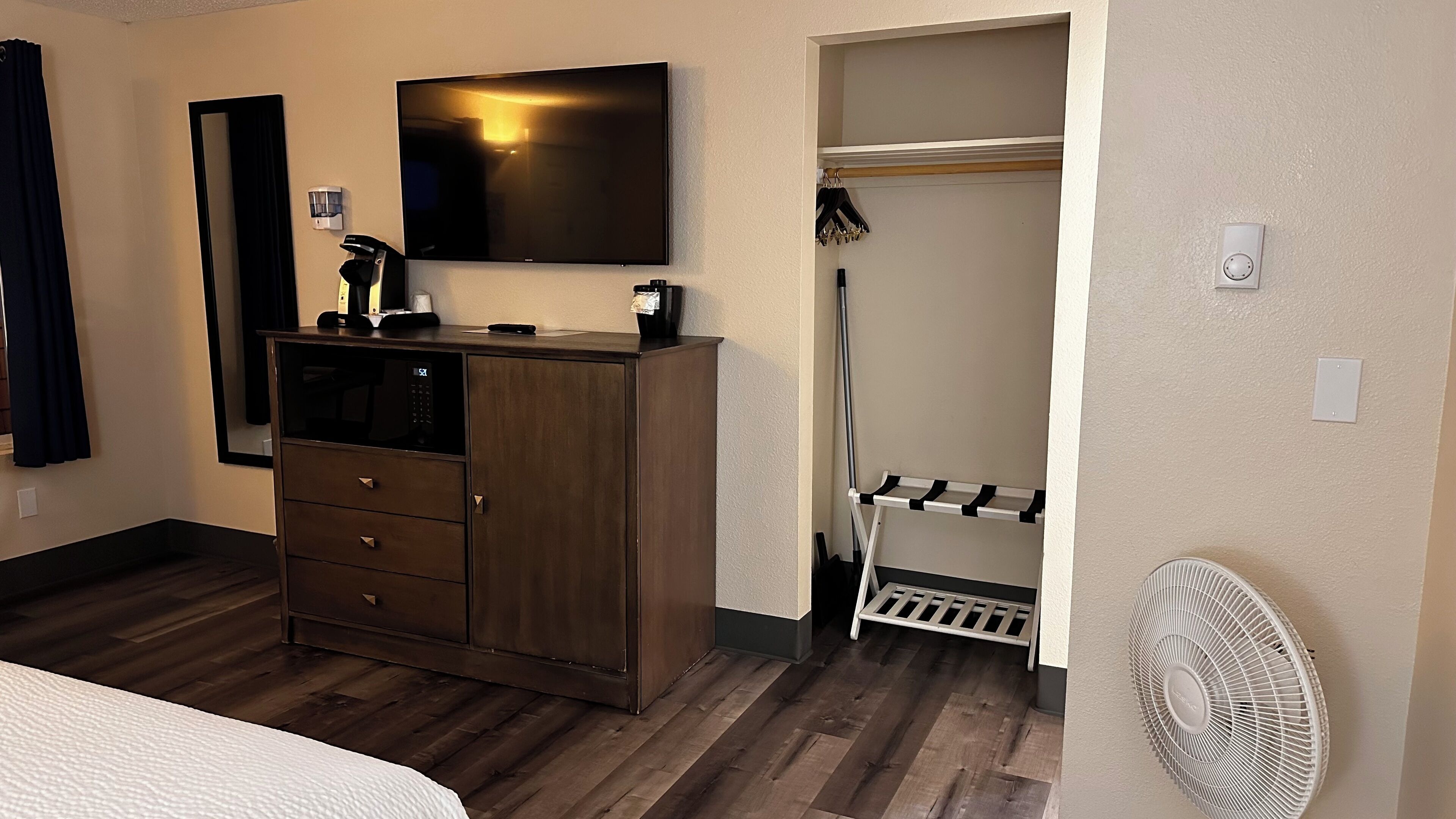 A hotel room with a wooden wardrobe, a flat-screen TV, a mirror, a coat rack, and a white fan.