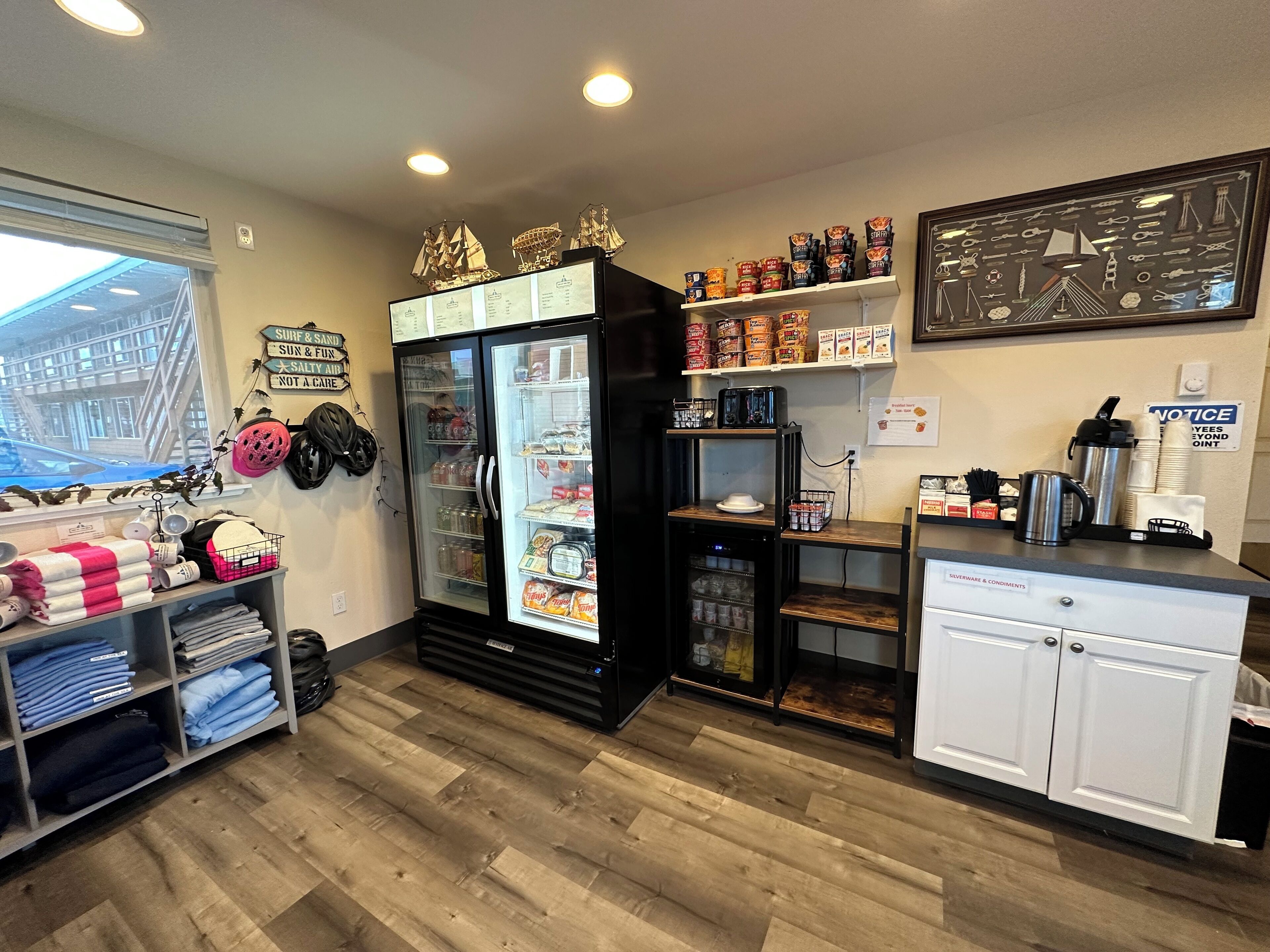 A kitchen area with a refrigerator, shelves with food items, a coffee machine, and a window with a view of a building outside.