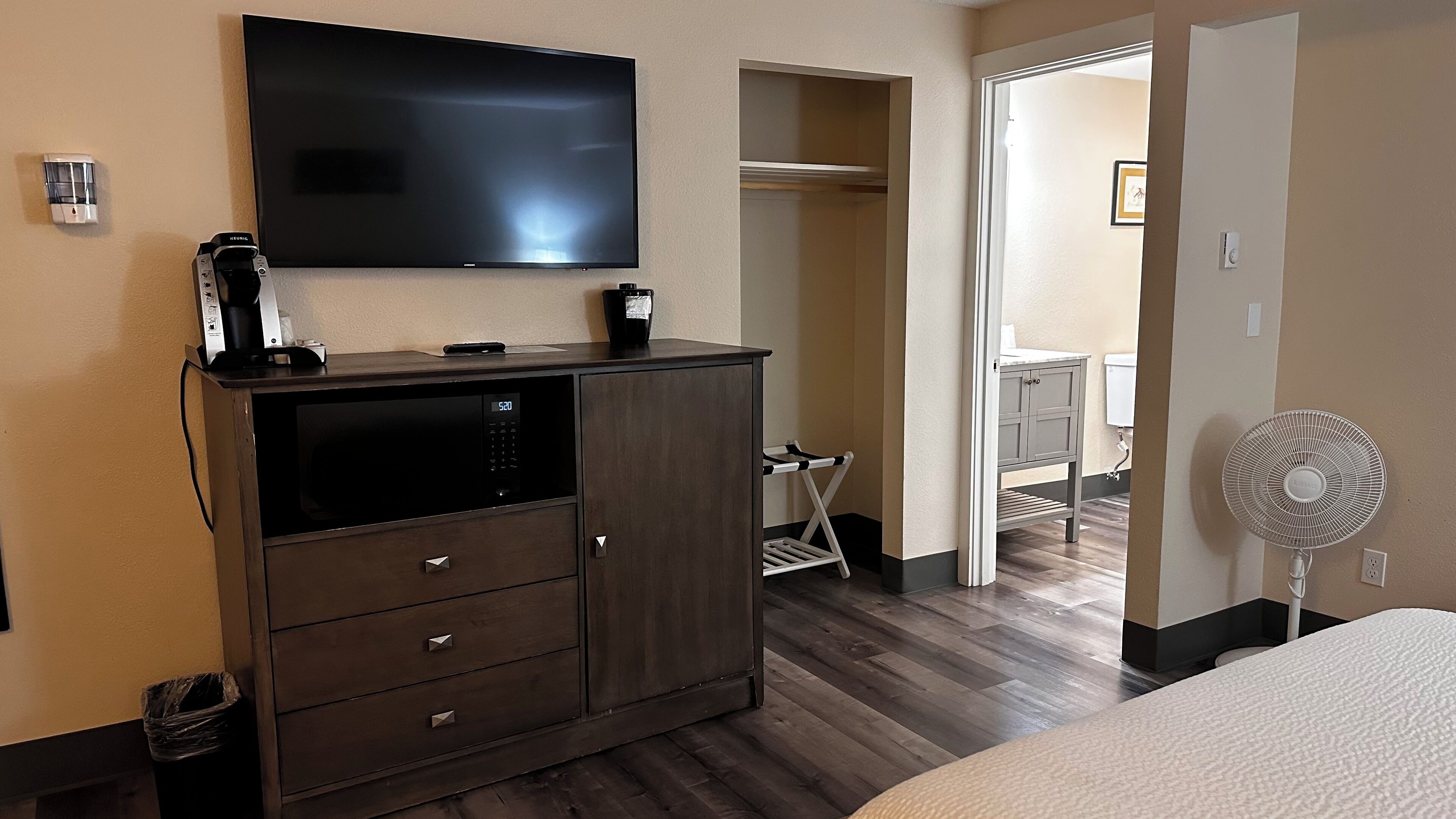 A hotel room with a large flat-screen TV mounted on the wall, a minibar, a wooden cabinet with drawers, a small table with a coffee maker, a white towel rack, a grey cabinet, and a standing fan.