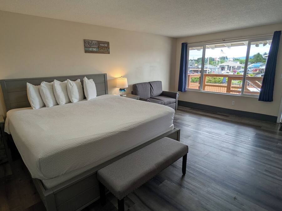 A hotel room with a large bed, a bench, a nightstand, a lamp, and a view of the outside.
