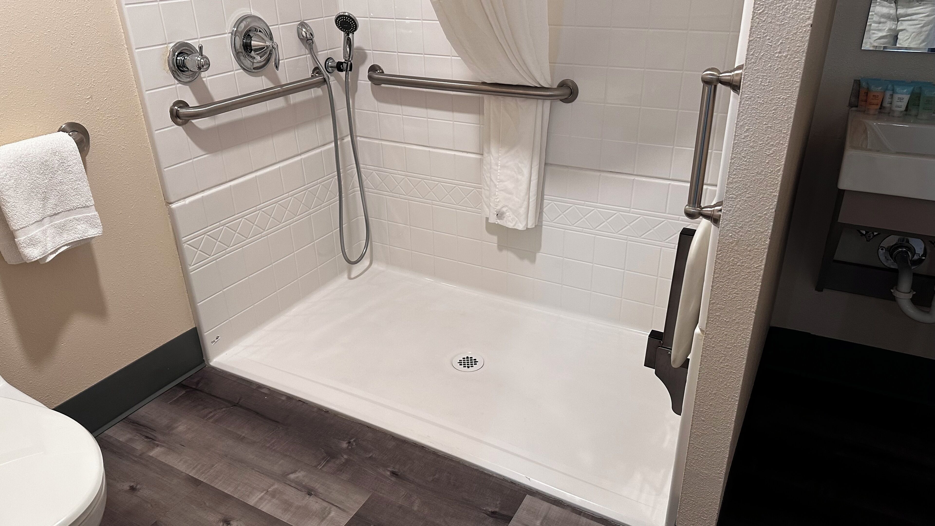 A walk-in shower with a grab bar, white tiled walls, and a wooden floor.