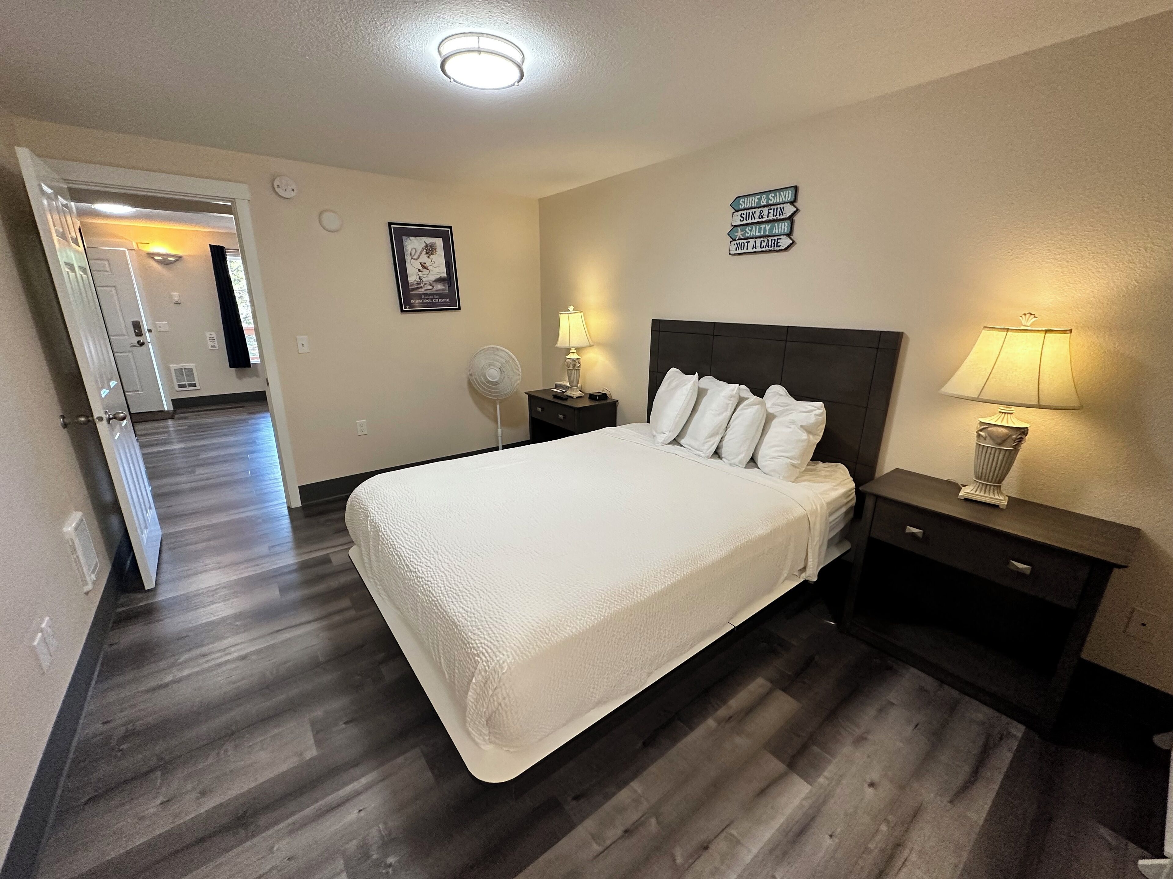 Una habitación de hotel con una cama grande, dos mesitas de noche con lámparas, un ventilador de pared y un cartel con texto.