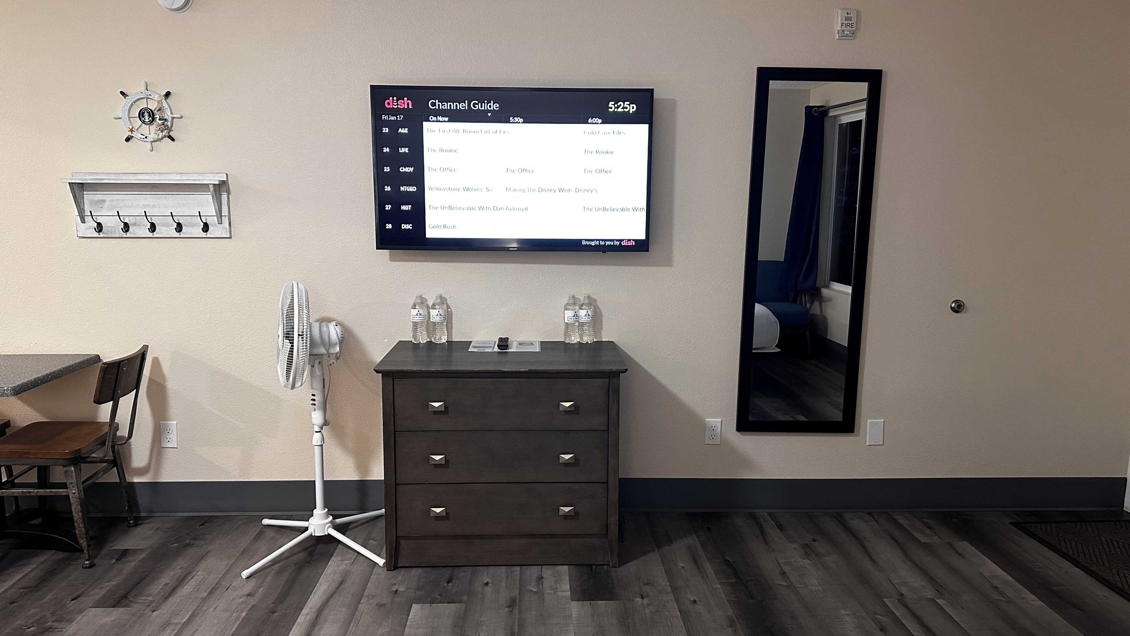 A hotel room with a flat-screen TV displaying a channel guide, a wooden dresser, a desk with a chair, a standing fan, and a mirror on the wall.