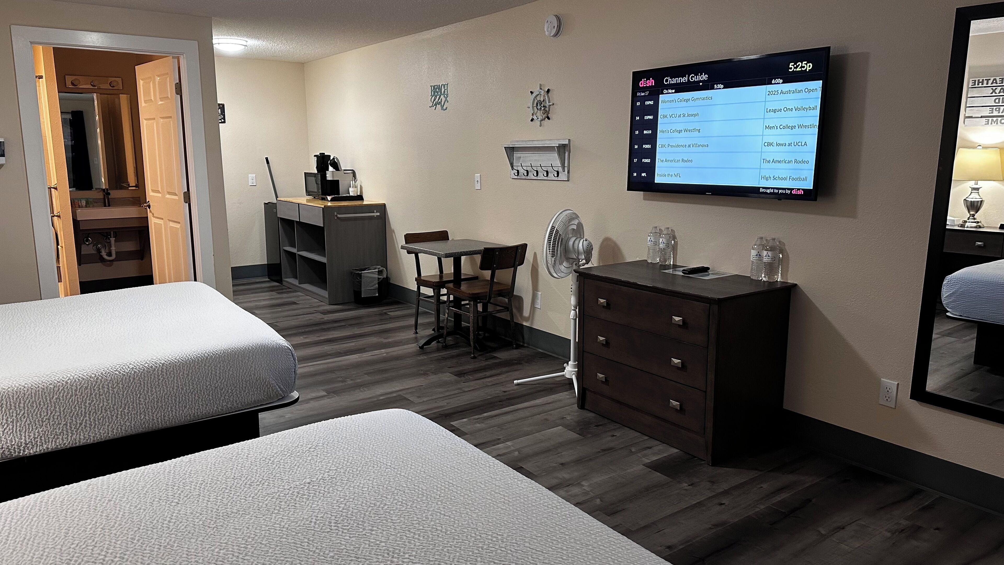 Habitación de hotel con dos camas, un escritorio, un aparador y un ventilador. En la televisión se muestra una guía de canales.
