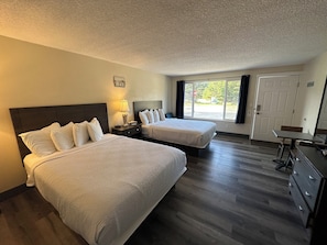 A hotel room with two beds, a desk, a dresser, a lamp, and a window with a view of the outdoors.