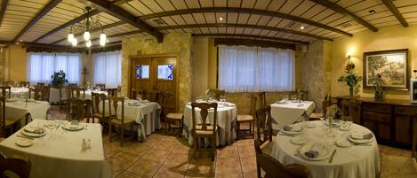 Un restaurante con varias mesas redondas, cada una con mantel blanco, platos y copas de vino. El espacio tiene paredes de piedra, vigas de madera en el techo y ventanas grandes con cortinas blancas.