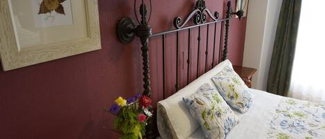 Un lit bien fait, avec une literie à motifs floraux, une table de chevet ornée d’un vase de fleurs et deux lampes fixées au mur.