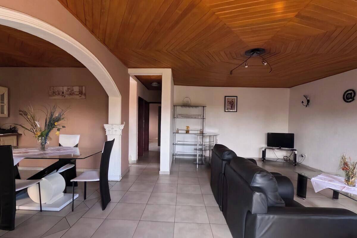 Un salon moderne avec un plafond en bois, un coin repas avec une table et des chaises blanches, un canapé en cuir noir et un téléviseur posé sur un meuble.