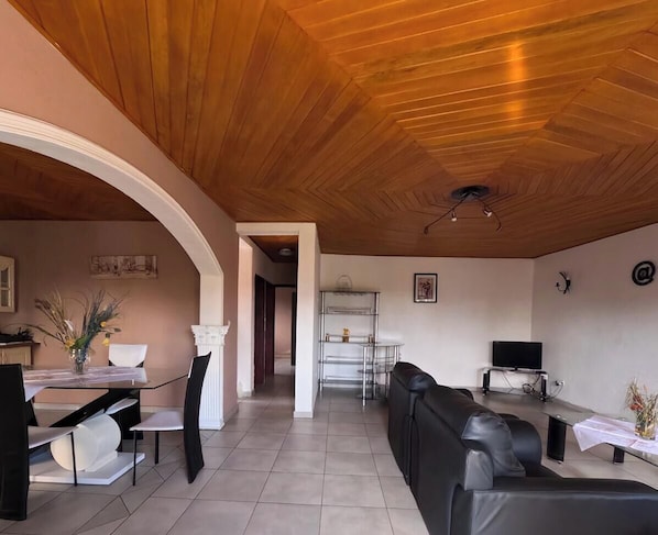 Un saló modern amb sostre de fusta, una zona de menjador amb taula i cadires blanques, un sofà de cuir negre i un televisor sobre un suport.