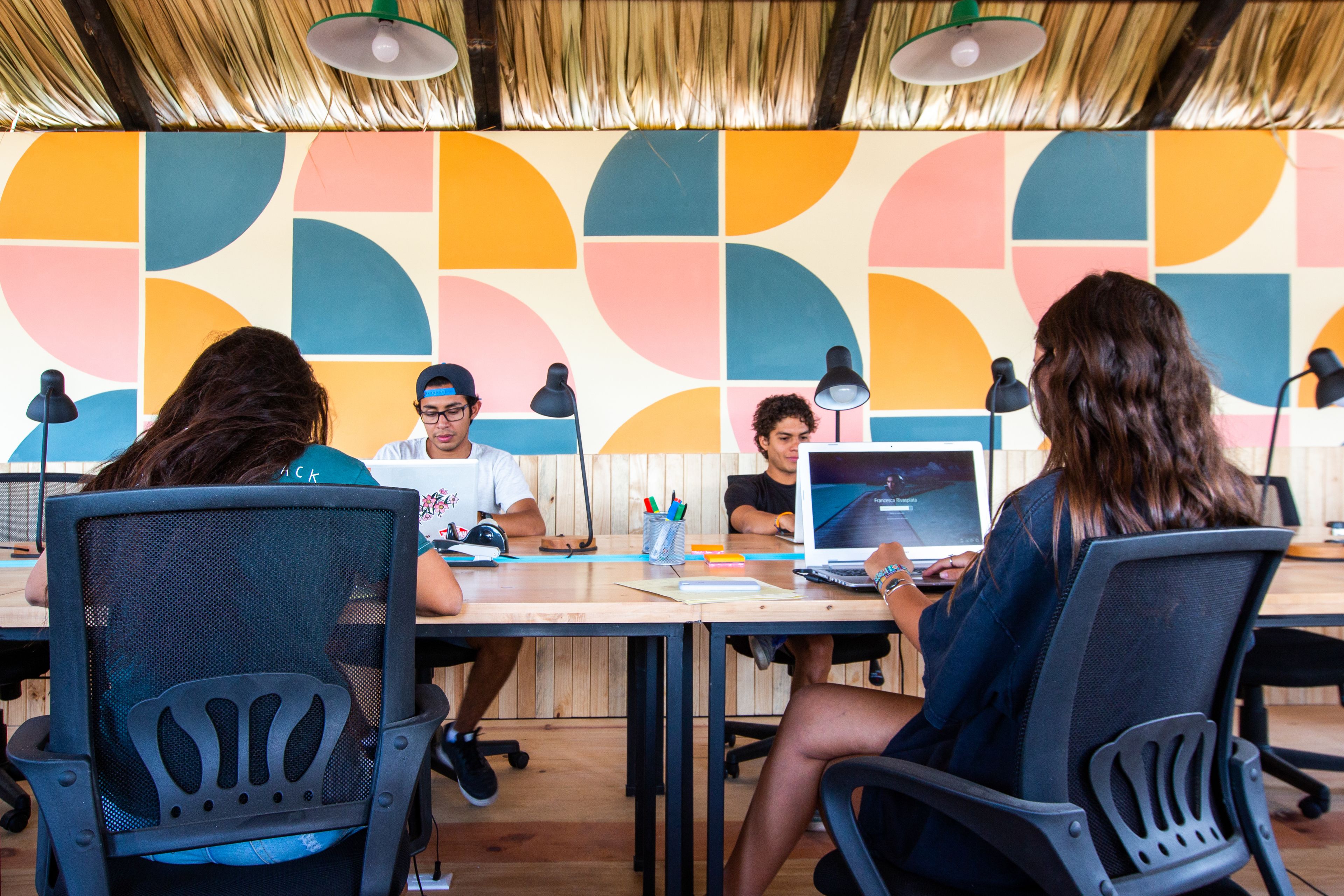 Un grupo de personas sentadas alrededor de una mesa con portátiles, en una sala con un llamativo diseño geométrico en la pared.